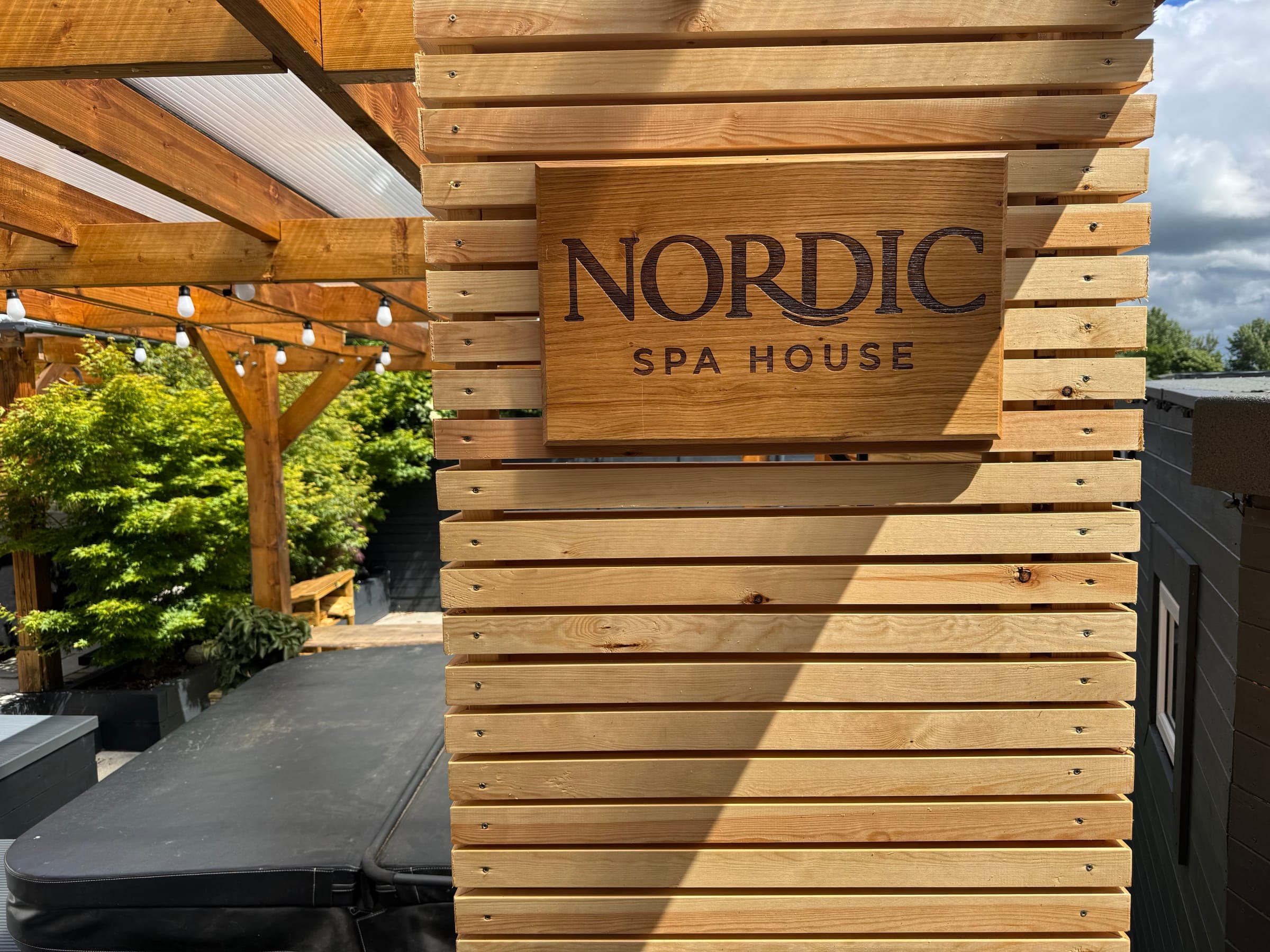 The carved Nordic Spa House sign on the cedar cladding at the garden entrance, beside the pergola