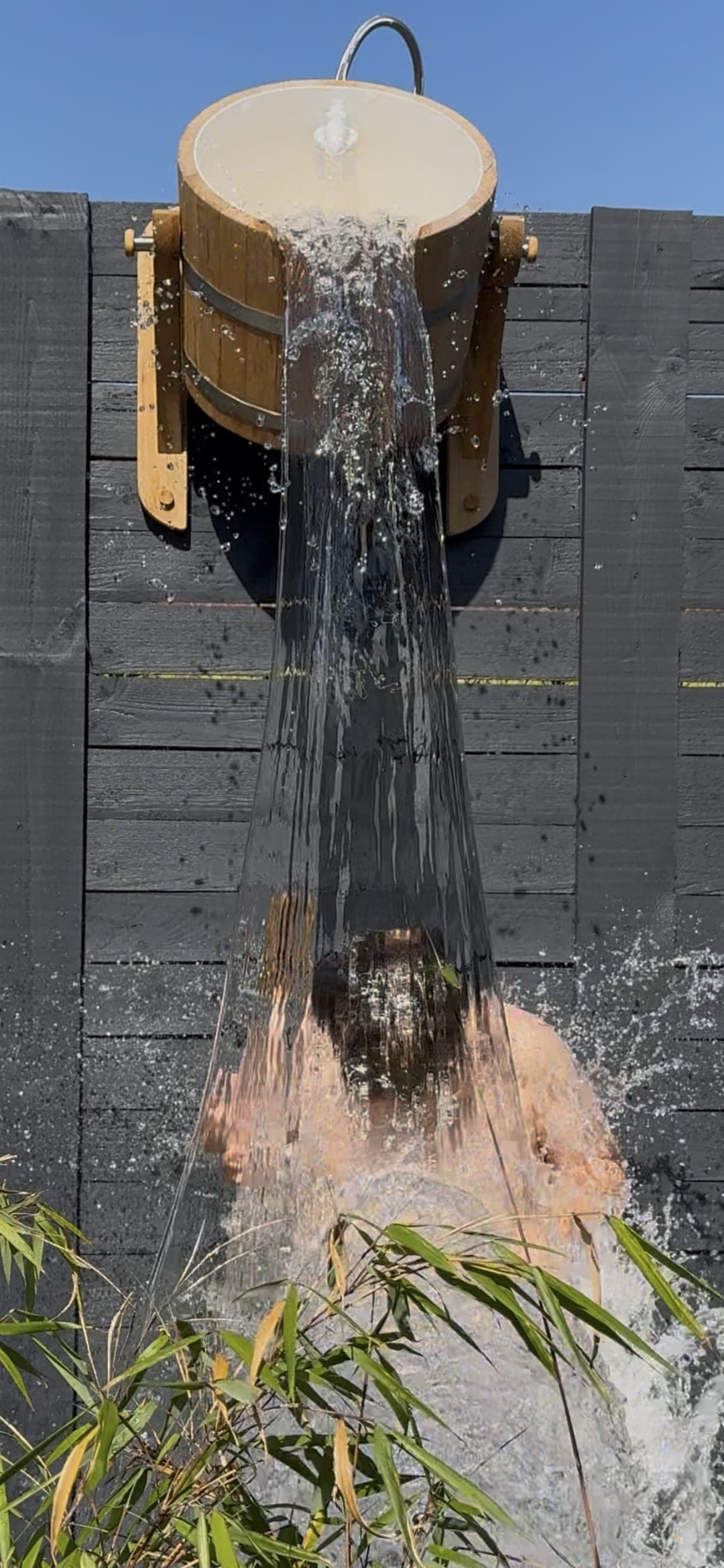 Cedar bucket tipping, a column of cold water pouring down the dark cladding past the bamboo